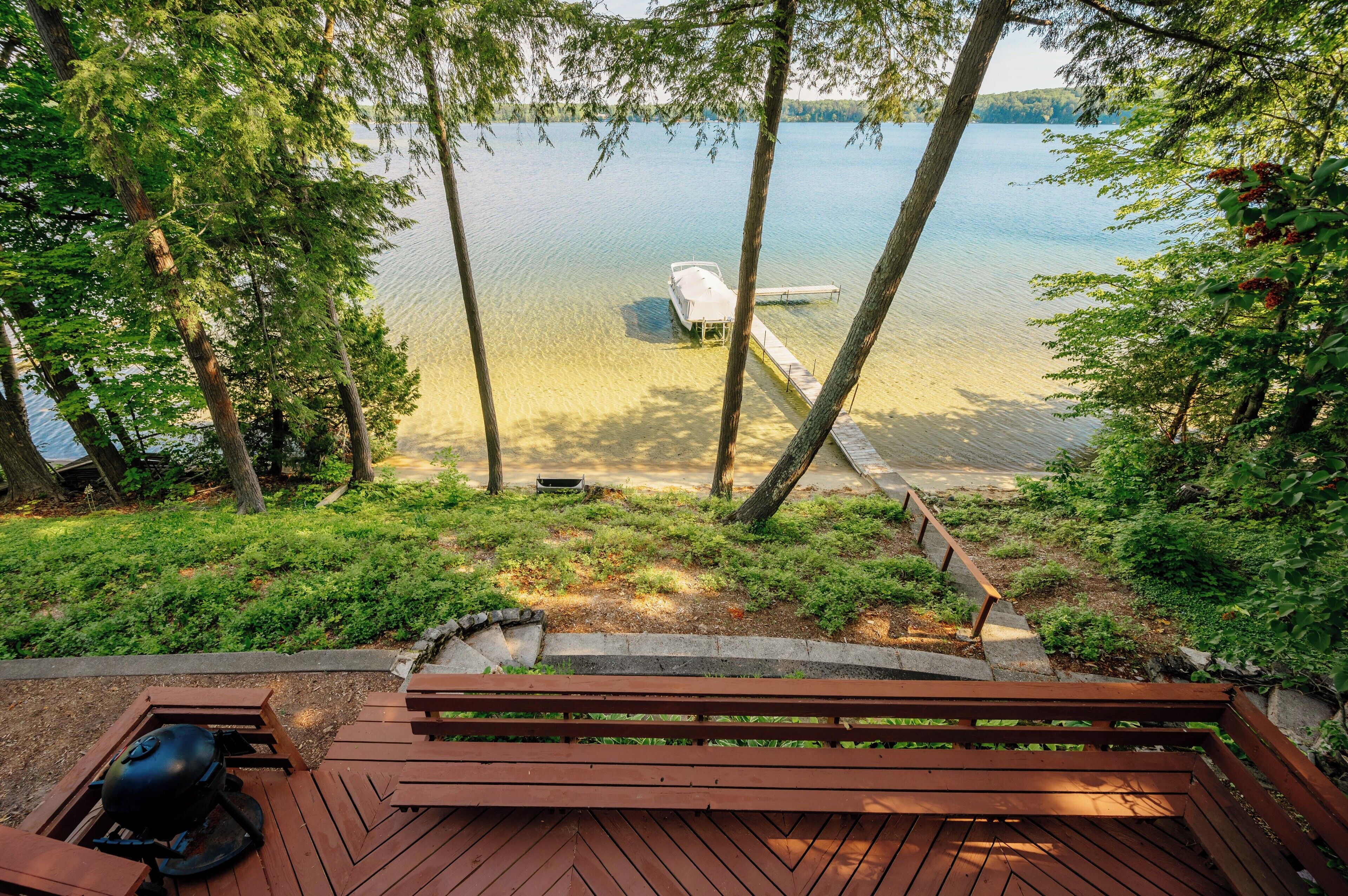 A wooden deck with a grill, overlooking a lake with a dock and boat.