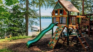 A wooden play structure with a slide and climbing wall, surrounded by trees and overlooking a body of water. - Spacious Waterfront Home on Long Lake (Traverse City)