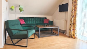 A living room with a green sofa, a green armchair, a coffee table, a floor lamp, and a TV mounted on the wall. - Ferienwohnung F18 - 2-zi. Fewo im Dachgeschoss für 3 Personen mit Wlan (Graal-Müritz)