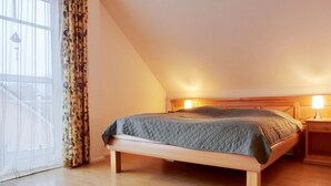 A bedroom with a wooden bed, a nightstand, a window with curtains, and a view of a balcony. - Ferienwohnung F18 - 2-zi. Fewo im Dachgeschoss für 3 Personen mit Wlan (Graal-Müritz)