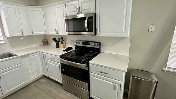 A kitchen with white cabinets, a stainless steel stove, a microwave, and a trash can.