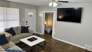 A living room with a beige sofa, a coffee table, a ceiling fan, a TV, and a door leading to another room.