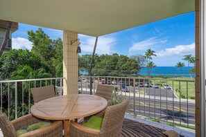 Un balcon avec une table ronde en bois et des chaises en osier, donnant sur une rue animée où l’on peut voir des voitures et des palmiers, et offrant une vue sur l’océan.