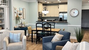 A modern living room with a dining area, kitchen, and a clock on the wall. - Windswept Dr  Condo in a quiet, golf course community close to Rehoboth Beach (Millsboro)