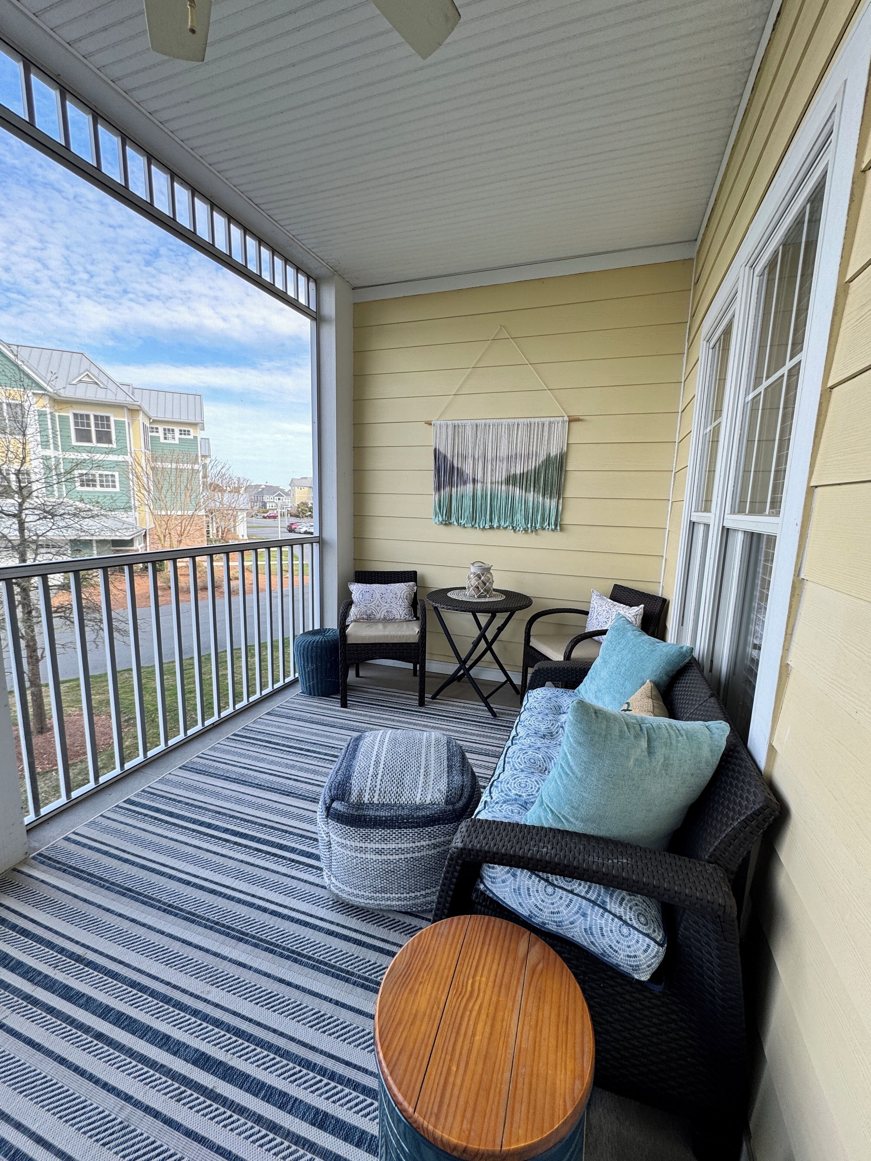A covered balcony with a striped rug, a wooden table, and a black sofa with cushions.