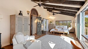 A spacious bedroom with a large bed, a sitting area with a white sofa, a wooden cabinet, and a large window offering a view of the outdoors. - Spacious Mesa Home with Great Outdoor Patio and Ocean Views! (Santa Barbara)