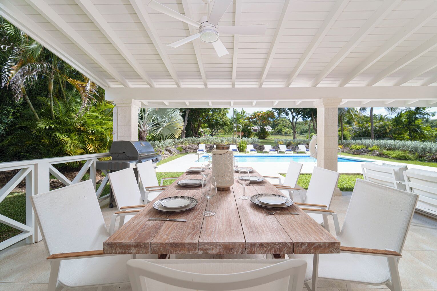 An outdoor patio with a wooden table set for dining, surrounded by white chairs, a ceiling fan, and a view of a swimming pool.