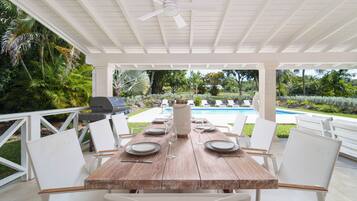 An outdoor patio with a wooden table set for dining, surrounded by white chairs, a ceiling fan, and a view of a swimming pool.
