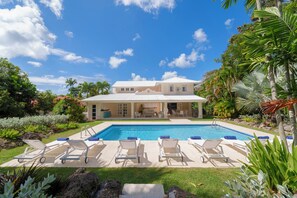 Ein zweigeschossiges Haus mit Swimmingpool, umgeben von einem Garten mit verschiedenen Pflanzen und Palmen.