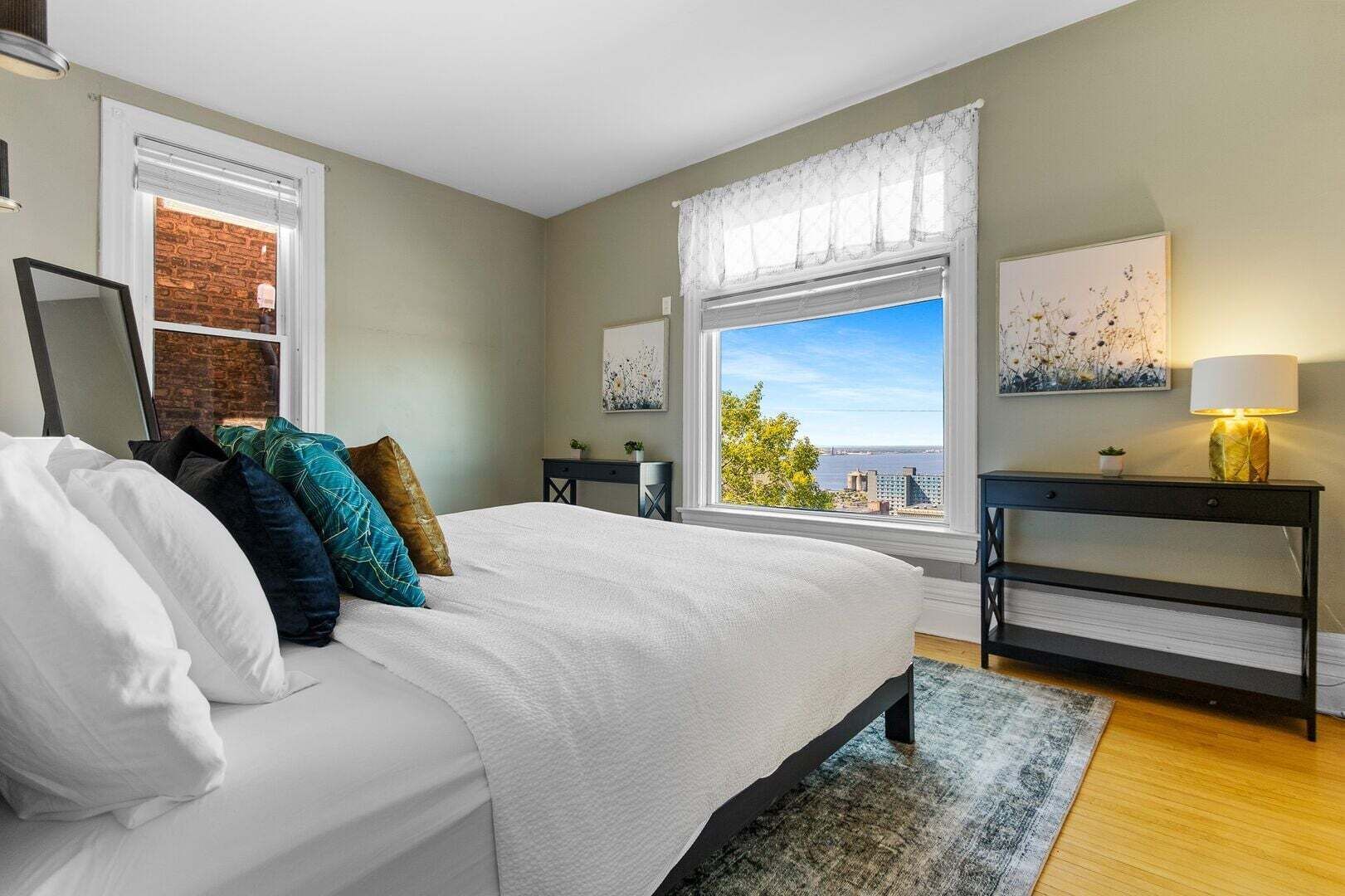 Un dormitorio con cama, una mesita de noche, una lámpara, un ventanal con vistas y un cuadro en la pared.