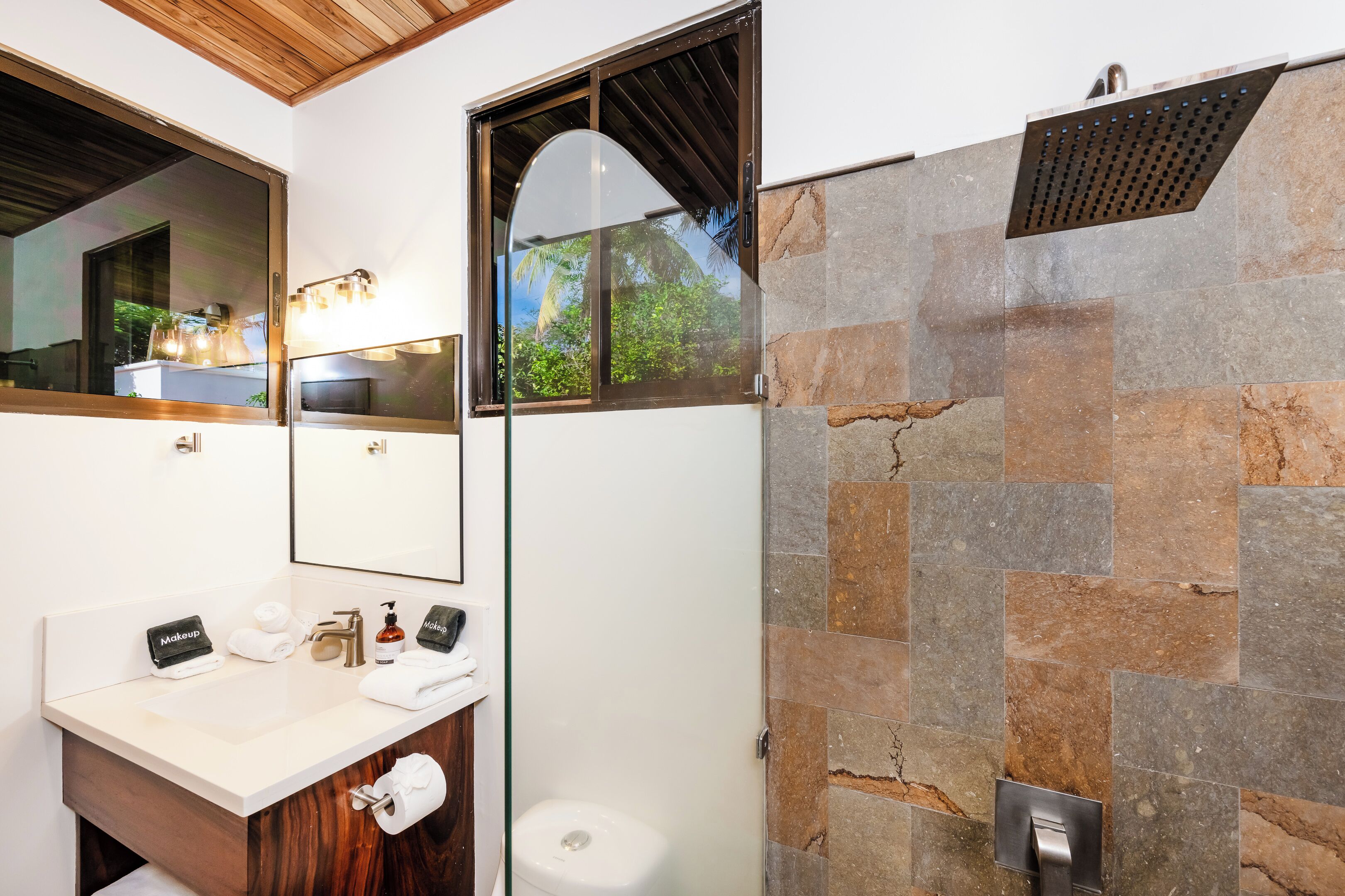A bathroom with a white sink, a mirror, a shower with stone tiles, and a window with a view of greenery.