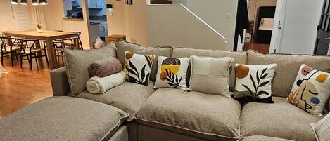 A modern living room with a sectional sofa, wooden flooring, a dining area, and a staircase leading to an upper level.