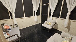 A room with a black wooden floor, white walls, and a white ceiling. There are two windows with white curtains, a white chair with a patterned cushion, and a white sofa with a patterned cushion. - Private Raleigh Bungalow Retreat near Downtown Raleigh (Raleigh)