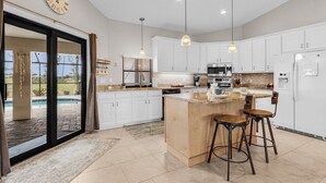 A modern kitchen with white cabinets, a large island with bar stools, a built-in oven, and a large window overlooking a pool. - Golf Course View! Heated Pool - Villa Fairway to Florida - Roelens Vacations (Cape Coral)