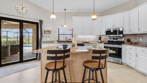 A modern kitchen with white cabinets, a central island with stools, a large window, and a view of a patio area. - Golf Course View! Heated Pool - Villa Fairway to Florida - Roelens Vacations (Cape Coral)