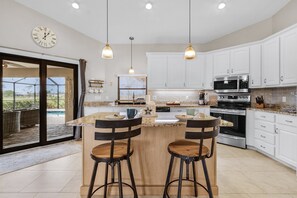 A modern kitchen with white cabinets, a central island with stools, a large window, and a view of a patio area. - Golf Course View! Heated Pool - Villa Fairway to Florida - Roelens Vacations (Cape Coral)
