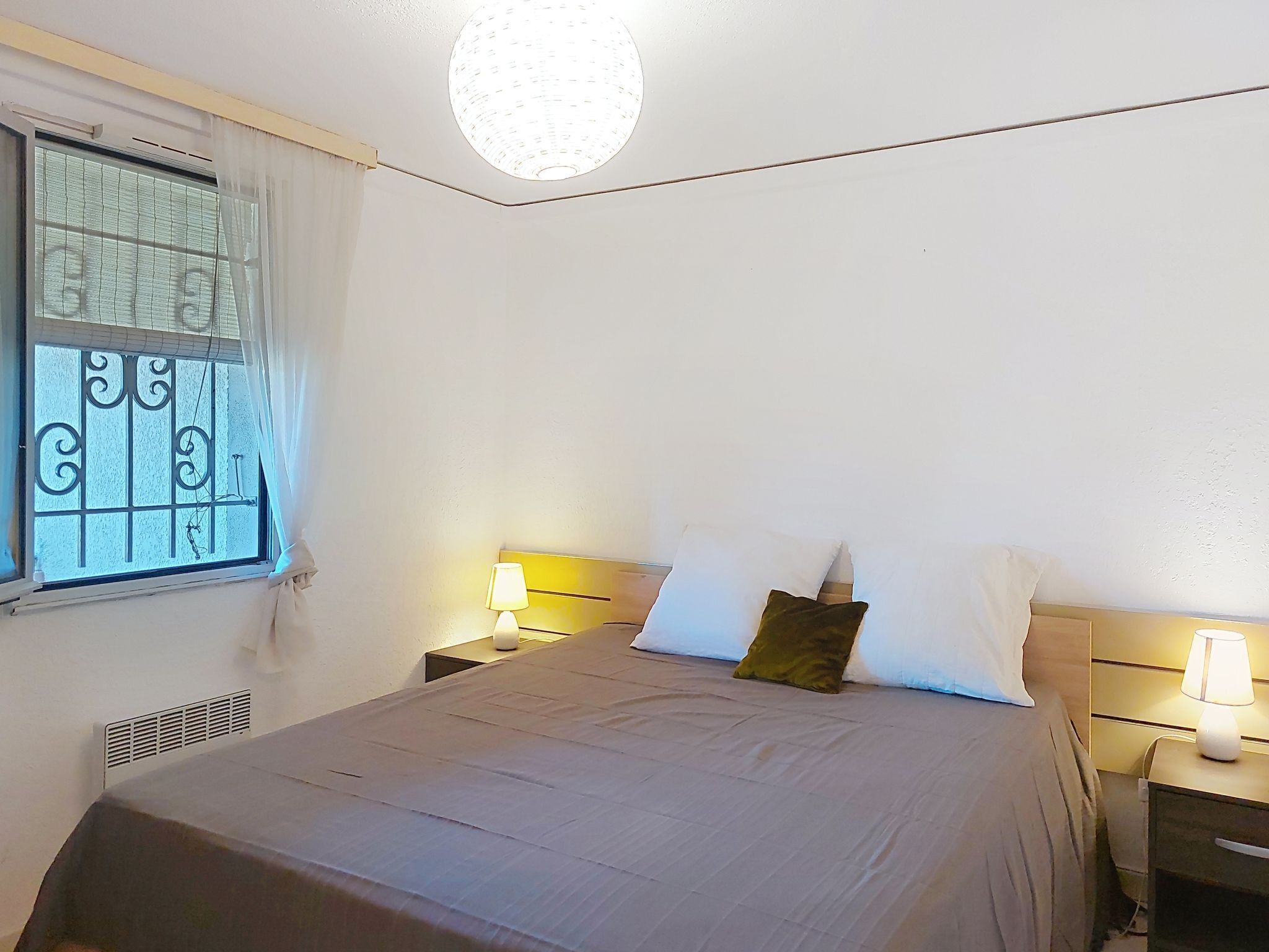 Une chambre à coucher avec un lit, deux tables de chevet avec des lampes, une fenêtre avec des barreaux en métal et un luminaire au plafond.