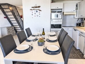 A dining area with a table set for four, a kitchen in the background, and a staircase leading to another level.