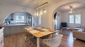 A spacious living area with a wooden dining table, white chairs, and a sofa. There is a kitchen in the background with grey cabinets and a window. - Casa Velazquez (Ciudad Quesada) (Rojales)