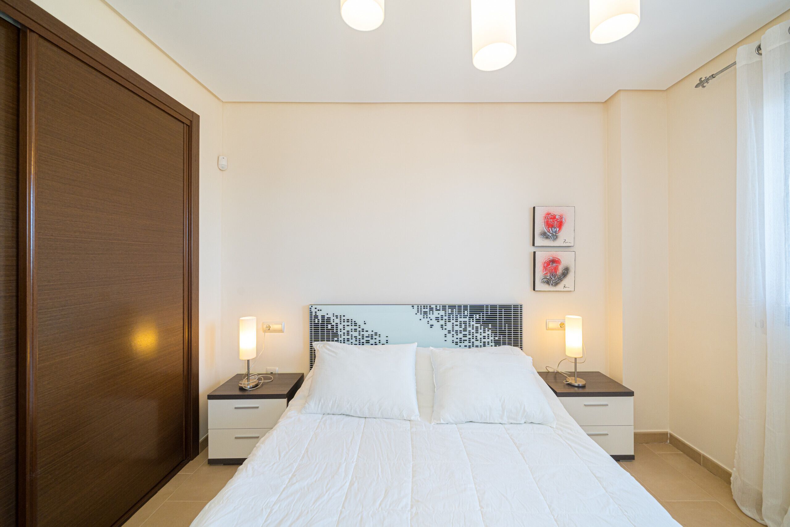 Ein Schlafzimmer mit einem großen Bett, zwei Nachttischen inklusive Lampen, einem Holzschrank und zwei gerahmten Bildern an der Wand.