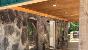 A covered outdoor seating area with stone walls, a wooden ceiling, and a view of a barbecue grill outside.