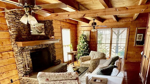 Interno accogliente di una baita con camino in pietra, travi a vista in legno, ventilatore a soffitto, albero di Natale e mensola del camino.