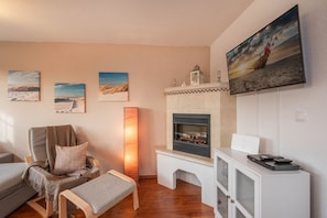 A living room with a fireplace, a mounted TV, and a painting of a beach scene. - Strandbrise - Ferienwohnung Strandbrise (Koserow)