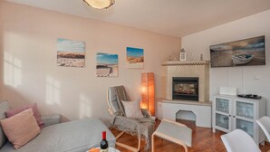 Una sala de estar acogedora con sofá, sillón y una pequeña mesa con botella de vino y copas. Hay una chimenea, televisor y arte mural con paisajes.