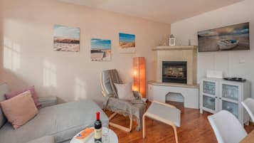 Un salon chaleureux avec un canapé, un fauteuil et une petite table sur laquelle se trouvent une bouteille de vin et des verres. On y trouve également une cheminée, un téléviseur et des œuvres d’art murales représentant des paysages.