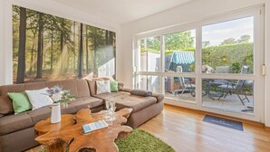 A living room with a large window, a brown sofa, a wooden coffee table, and a forest mural. - Reni - Idyllisches Erdgeschoss-appartement Terrasse & Strandnah (Koserow)