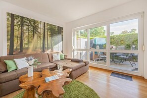 Un salon doté d’une grande fenêtre, d’un canapé marron, d’une table basse en bois et d’une fresque représentant une forêt.