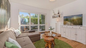 A modern living room with a sectional sofa, a wooden coffee table, a TV, and a large window with a view of greenery. - Reni - Idyllisches Erdgeschoss-appartement Terrasse & Strandnah (Koserow)
