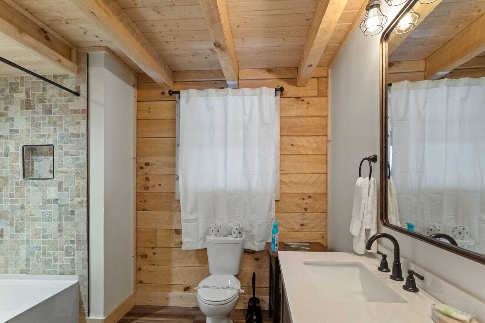 Kylpyhuone, jossa on puinen katto, laatoitettu suihkuseinä, wc-istuin, peilillä varustettu pesuallas ja pyyheteline.