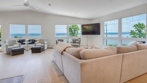 Amplia sala de estar con un sofá beige grande, dos sillones, una mesita de centro y un televisor de pantalla plana montado en la pared. Amplias ventanas ofrecen vista al mar y a un muelle.
