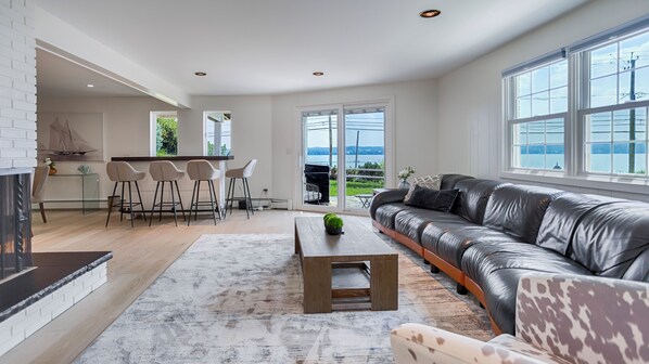 A spacious living area with a leather sofa, a coffee table, and a fireplace. There is a kitchen area with bar stools and a view of the outdoors through large windows.