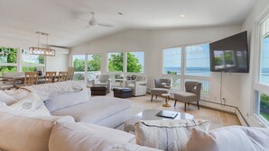 Un salón amplio con un sofá grande, varias sillas, una mesa de centro y un televisor de pantalla plana. Grandes ventanales ofrecen vistas al exterior.