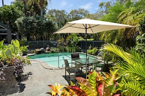 An outdoor swimming pool with a glass table and chairs, surrounded by lush greenery and a metal fence.