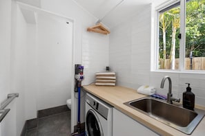 A compact kitchen with a washing machine, a sink, and a window overlooking greenery. - Villa Le Miramar Perfect for your Pooch Holiday (Noosa)