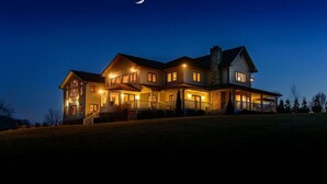 A large, two-story house with multiple windows and a covered porch, set on a grassy hill under a night sky with a crescent moon.