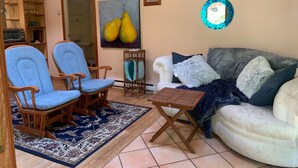 Una sala de estar con un sillón azul, una mesa de centro de madera, un sofá blanco con cojines azules y grises, un tapete estampado y un cuadro de peras en la pared.