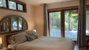 Un dormitorio con una cama grande, cabecera de madera, mesita de noche con lámpara y ventana con vistas a árboles y un estacionamiento.