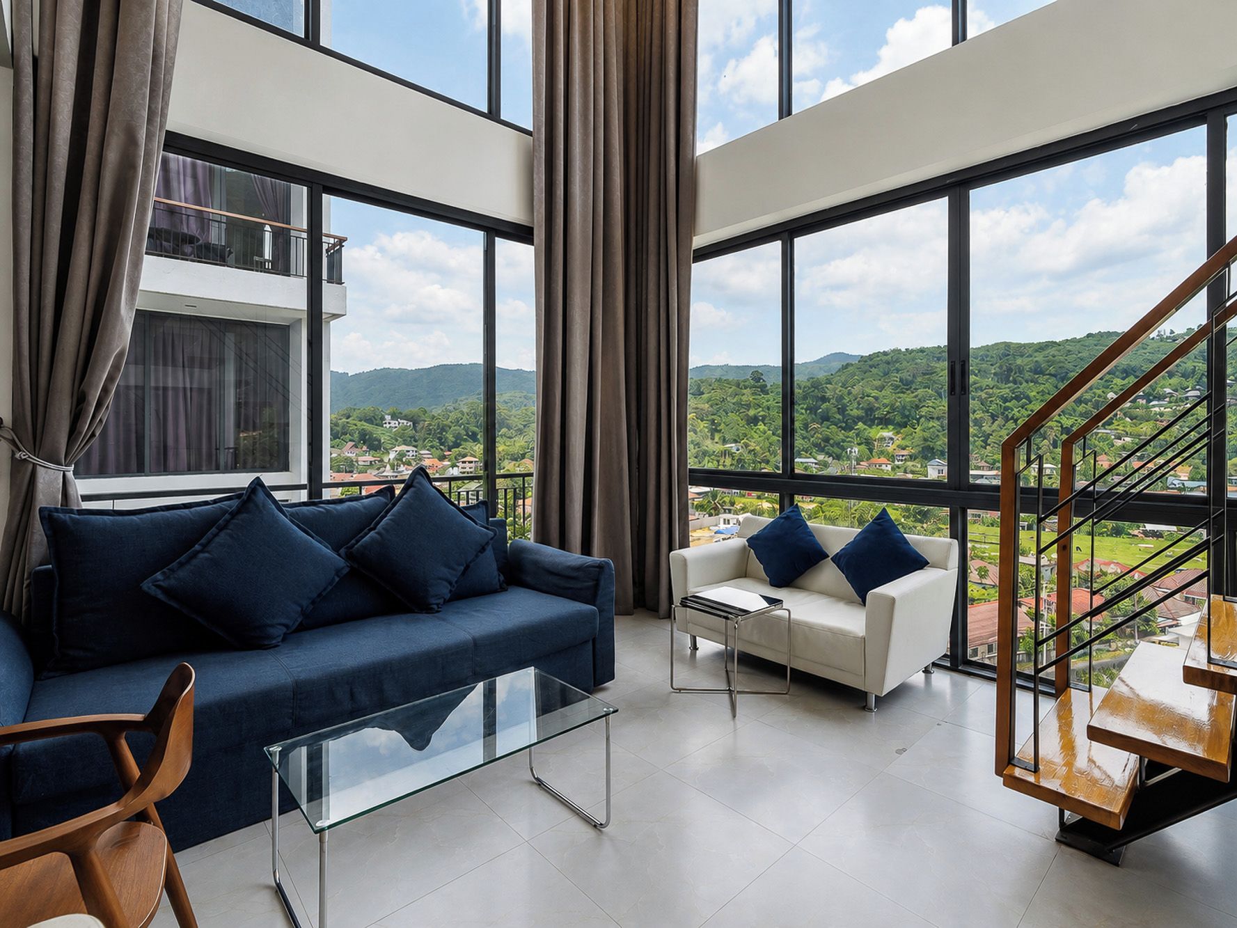 Una sala de estar moderna con una mesa de centro de vidrio, dos sofás con cojines azules y vistas a un paisaje verde a través de grandes ventanales.