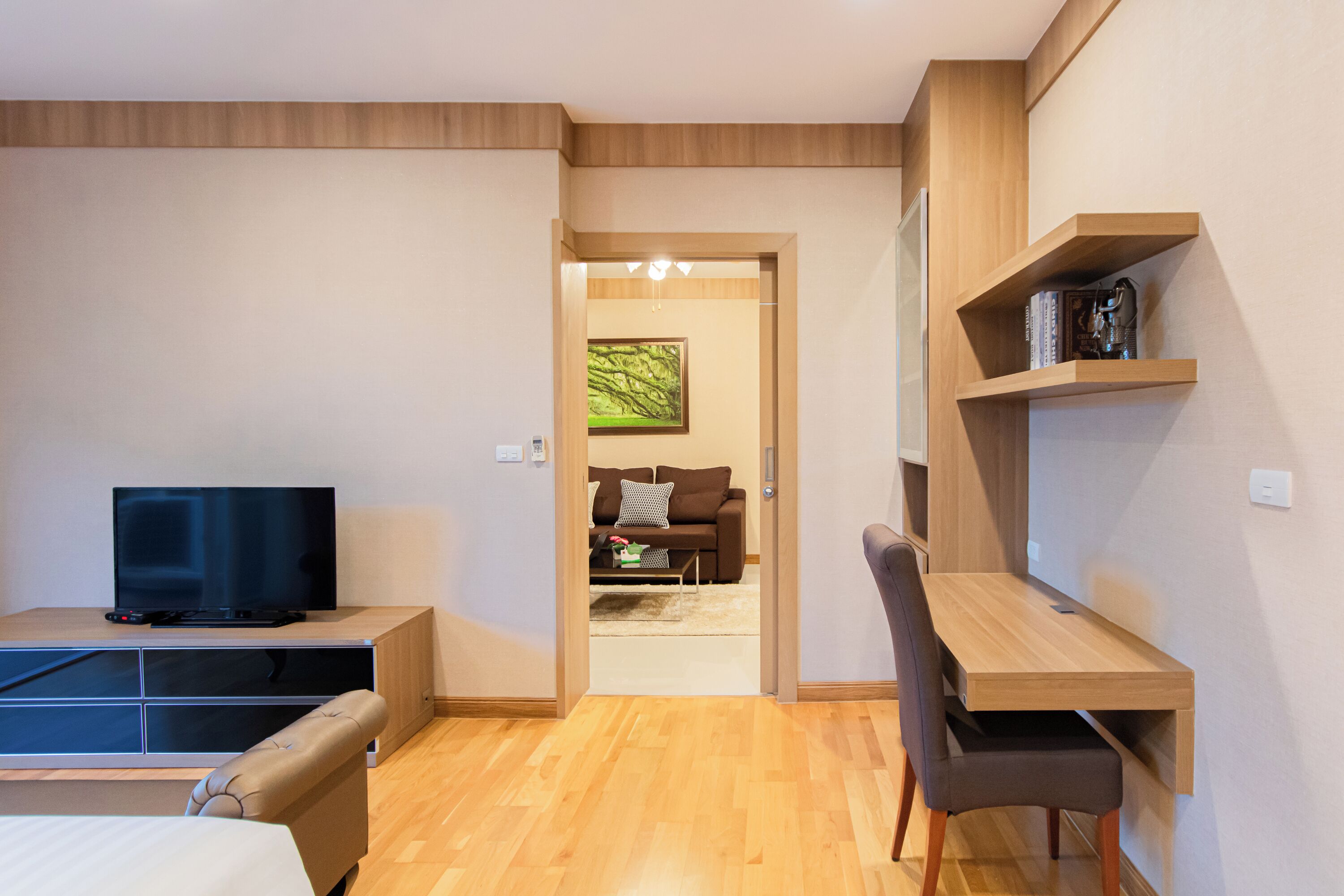 A modern hotel room with a wooden desk, a television, a sofa, and a bookshelf.