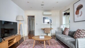 Ein modernes Wohnzimmer mit Flachbildfernseher, Couchtisch aus Holz, grauem Sofa mit gemusterten Kissen, Stehlampe und einem gerahmten Kunstwerk an der Wand.