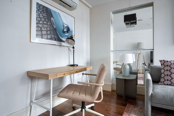 Una habitación moderna y luminosa con un escritorio de madera, una silla acolchada, una mesa de vidrio, un sofá gris y un cuadro en la pared.