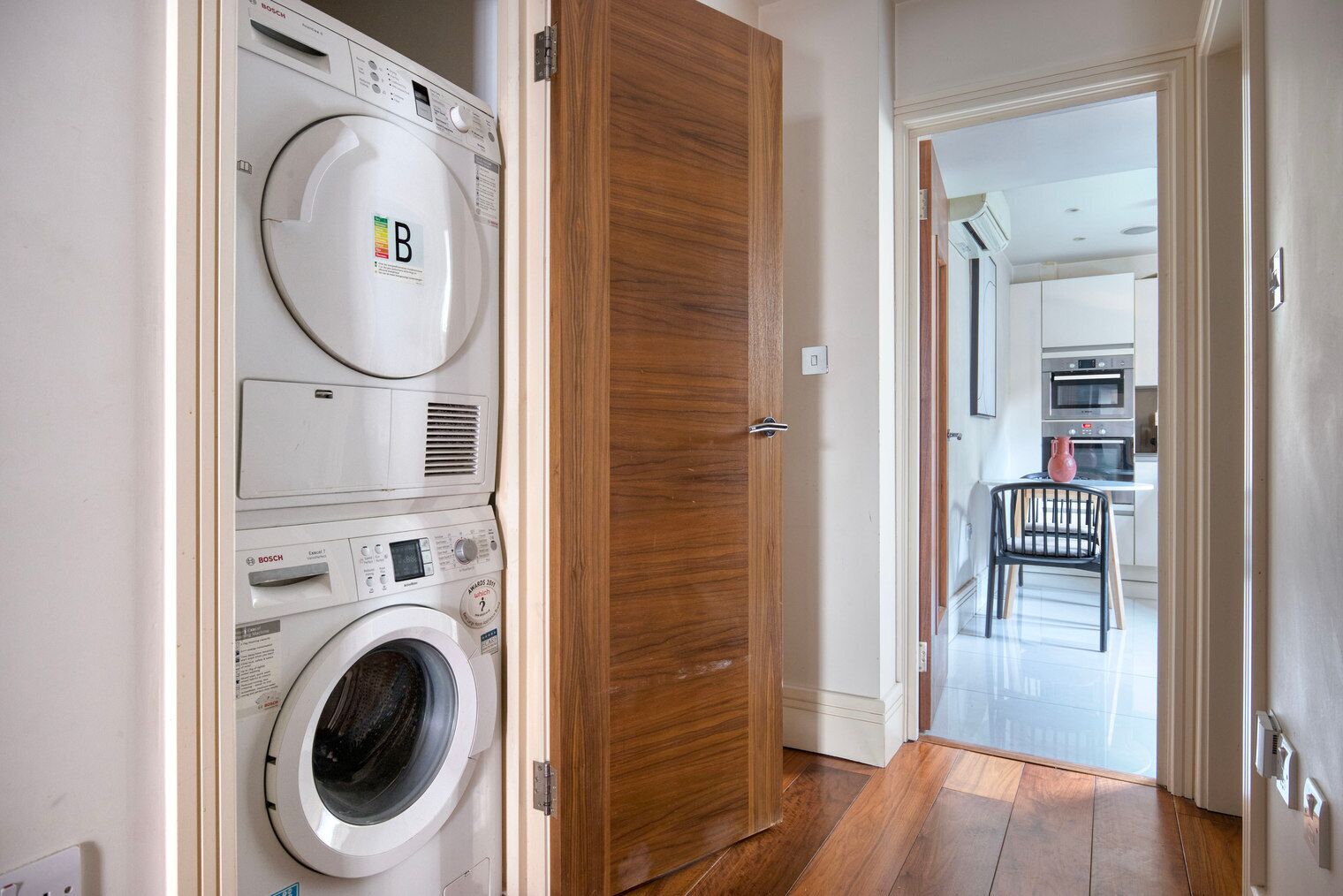 A laundry room with stacked washing machines and dryers, a wooden door, and a view into a modern kitchen with a dining area.