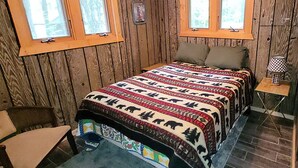 Una habitación de cabaña de madera con una cama, dos ventanas, una mesita y una silla.