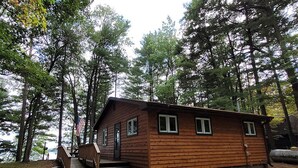 A small wooden cabin with a porch and a flag, surrounded by tall trees.
