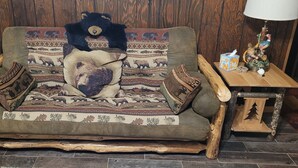 Un sofá de madera rústica con cojines de motivos animales, una mesita de madera pequeña con una lámpara y objetos decorativos, y una manta con estampado.
