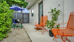 An outdoor patio with a barbecue grill, two orange chairs, a blue umbrella, and a potted plant.